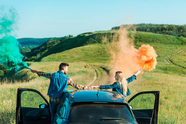 rear view of couple with colorful bombs standing on car and holding hands on rural meadow - Фото, зображення