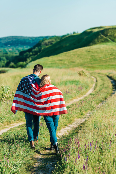 Rückansicht von jungem Paar mit amerikanischer Flagge auf ländlicher Wiese, Konzept zum Unabhängigkeitstag - Foto, Bild