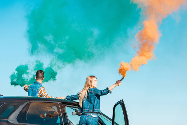 young couple standing on car and holding green and orange smoke bombs  - Photo, Image