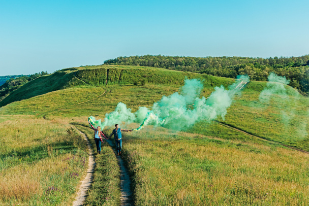 Weite Sicht auf junges Paar mit grünen Rauchbomben auf bäuerlicher Wiese  - Foto, Bild
