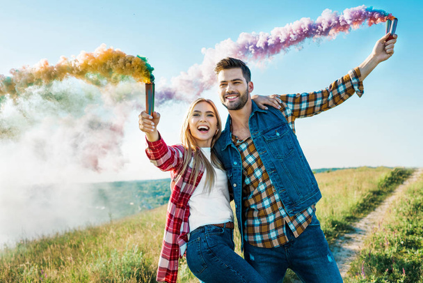 smiling couple holding colorful smoke bombs on rural meadow  - Zdjęcie, obraz