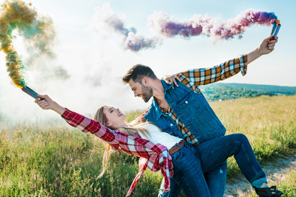side view of man with colorful smoke bombs holding girlfriend back in rural field  - Foto, Imagem