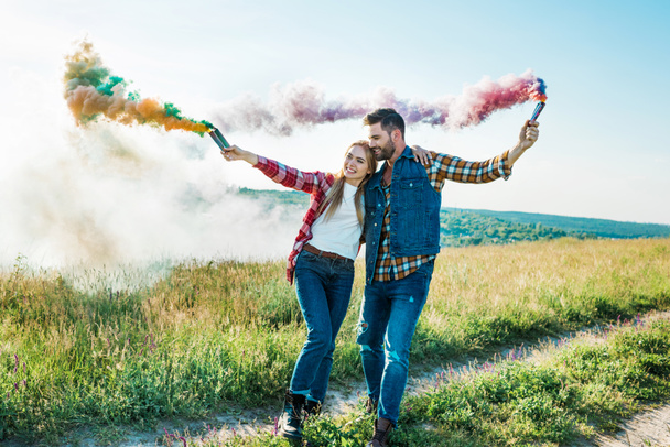 young couple holding colorful smoke bombs in field  - Фото, зображення
