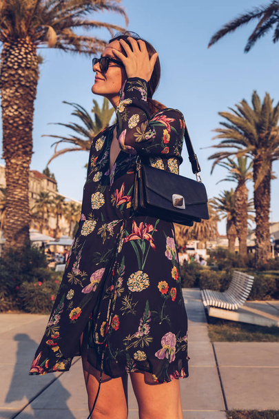 приваблива жінка позує на відкритому повітрі в літній день. блогер моди в квітковій міні-сукні, одягнений в сонцезахисні окуляри і тримає невелику чорну сумку. фон з пальмами
 - Фото, зображення