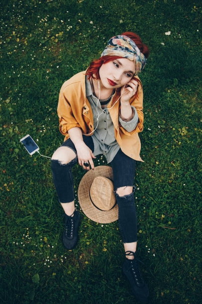 mooi meisje zittend op het gras en poseren met koptelefoon. modieuze gelukkige vrouw in trendy festival kleren genieten van het warme weer buiten, ontdekken van nieuwe muziek. Streetstyle - Foto, afbeelding