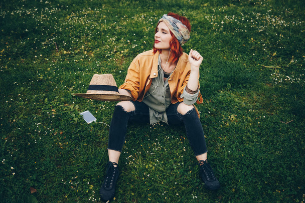 beautiful girl sitting on the grass and posing with headphones. fashionable happy woman in trendy festival clothes enjoying the warm weather outside, discovering new music. street style - Foto, Imagen