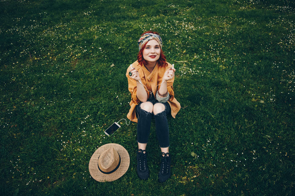 krásná dívka sedící v trávě a pózuje se sluchátky. Módní šťastná žena v módní festival oblečení těší teplé počasí venku, objevovat novou hudbu. pouliční styl - Fotografie, Obrázek