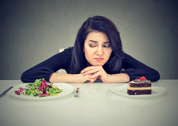 Beautiful brunette sitting at desk and suffering from choice between healthy salad and sweet dessert - Fotografie, Obrázek