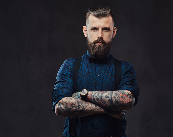 Portrait of a handsome old-fashioned hipster in a blue shirt and suspenders, standing with crossed arms in a studio. - Photo, Image