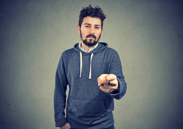 Bearded hipster in gray hoodie pointing at camera indicating with finger on gray background.  - Photo, Image