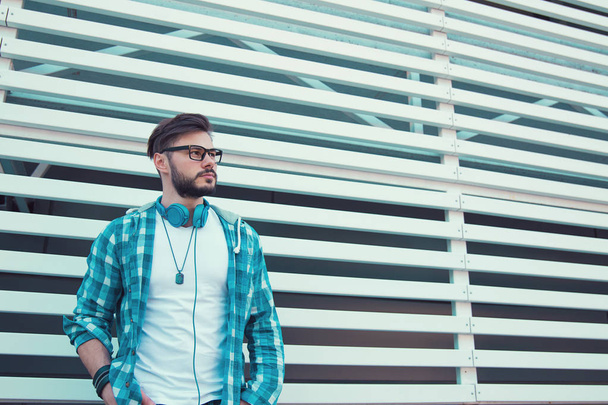 Trendy hipster man in glasses and blue shirt with headphones standing with hands in pockets looking away confidently - Photo, image