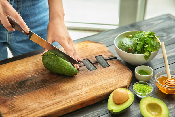 Jonge vrouw snijden avocado voor voedende masker aan tafel - Foto, afbeelding