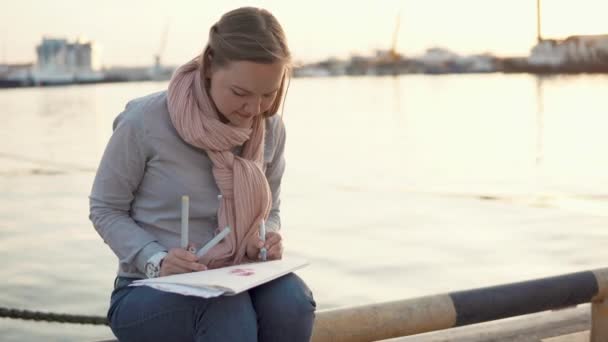 Młoda kobieta siedzi w pobliżu molo wiosną wieczorem i rysunek przez ołówki - Materiał filmowy, wideo
