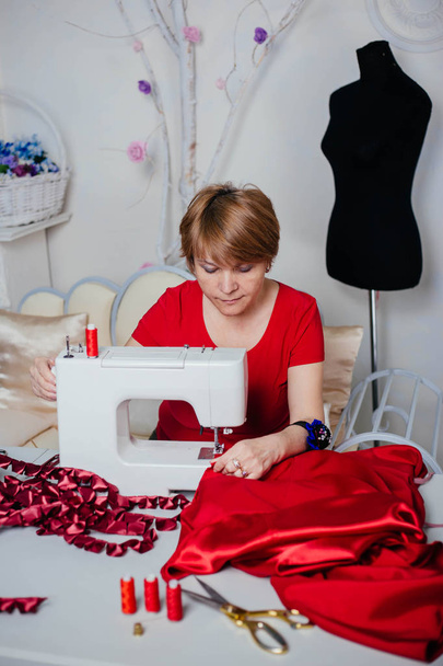 Eine Näherin in roter Kleidung näht an einer Nähmaschine. Atelier, der Prozess der Schneiderei. - Foto, Bild