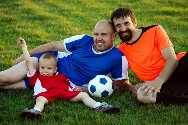 Отец, сын и дядя из футбольной семьи в футбольной спортивной форме лежат на траве в игровом поле и позируют. Счастливая семья в спортивной концепции
. - Фото, изображение