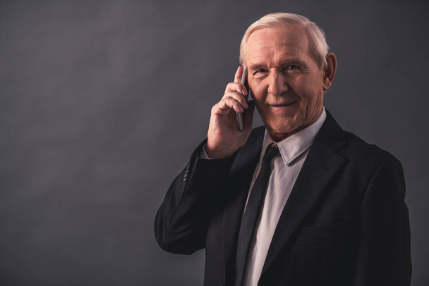 schöner alter Geschäftsmann im Anzug spricht auf dem Handy, blickt in die Kamera und lächelt, auf grauem Hintergrund - Foto, Bild