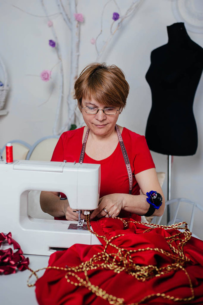 赤い服を着た女性の裁縫師の女性は、ミシンで縫います。アトリエ、仕立てのプロセス. - 写真・画像