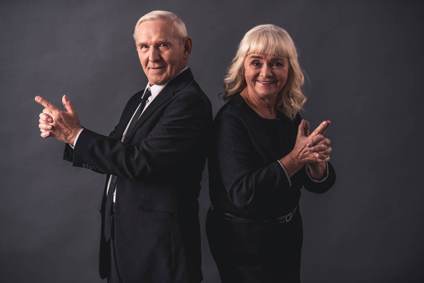 Beautiful old couple in elegant clothes is holding hands together imitating guns, looking at camera and smiling, on gray background - Фото, зображення
