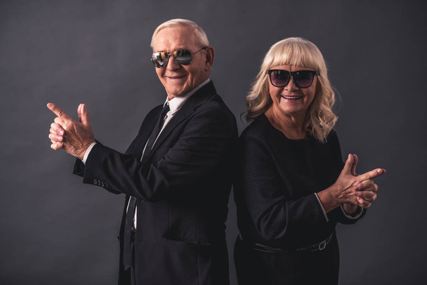 Hermosa pareja de edad en ropa elegante y gafas está sosteniendo las manos juntas imitando armas, mirando a la cámara y sonriendo, sobre un fondo gris
 - Foto, imagen