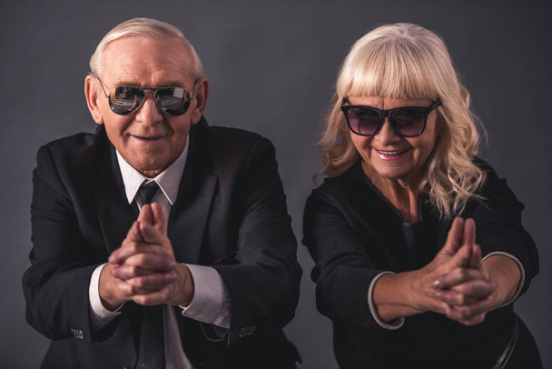 Beautiful old couple in elegant clothes and glasses is holding hands together imitating guns, looking at camera and smiling, on gray background - Photo, Image