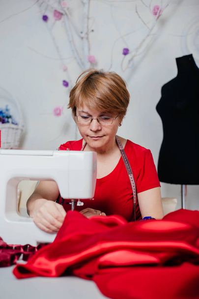 Eine Näherin in roter Kleidung näht an einer Nähmaschine. Atelier, der Prozess der Schneiderei. - Foto, Bild