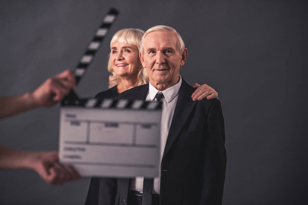 Hermoso anciano y mujer en ropa elegante sobre fondo gris, clapperboard en frente
 - Foto, imagen