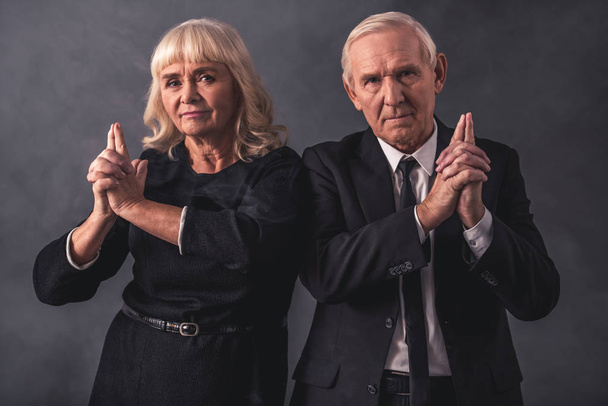 Krásný starý pár v elegantní oblečení je drží za ruce, napodobující zbraně, na šedém pozadí - Fotografie, Obrázek