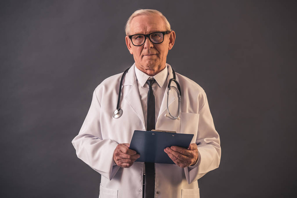Handsome old doctor in medical coat and eyeglasses is holding a folder and looking at camera, on gray background - Photo, Image