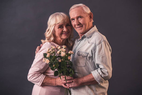 Hermoso anciano y mujer en ropa casual están sosteniendo flores, mirando a la cámara y sonriendo, sobre fondo gris
 - Foto, Imagen