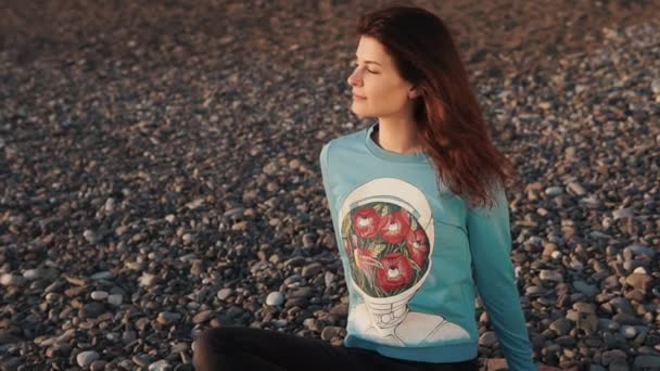 Χαρούμενη κοπέλα κάθεται σταυρωμένα πόδια σε μια παραλία με βότσαλα στο χρόνο βράδυ - Πλάνα, βίντεο