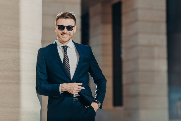 Позитивный молодой мужчина носит официальный костюм, носит модные оттенки, держит руку на кармане, противостоит офисному интерьеру, будучи в хорошем настроении после успешной деловой встречи. Концепция лидерства
 - Фото, изображение