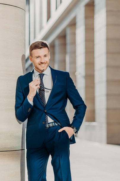 Thoughtful cheerful bearded male entrepreneur has pleasant smile, wears black suit, holds sunglasses, poses indoor, being delighted with successful business meeting and signing contract with partner - Foto, Imagen
