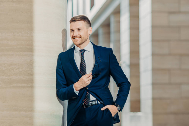 Image of successful businessman in elegant clothing, keeps hand in pocket, holds sunglasses, smiles cheerfully, looks somewhere, being confident in his business deal. People and job concept. - Photo, Image
