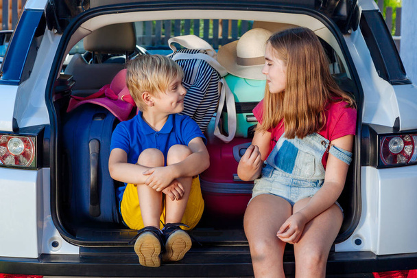 Маленька сестра і брат сидять у багажнику автомобіля з валізами. Подорож на машині сімейна поїздка разом відпустка. Сміється Дівчинка і хлопчик готові подорожувати з сім'єю
. - Фото, зображення
