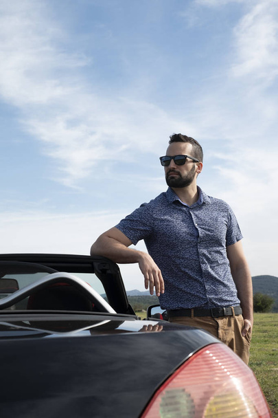Uomo con occhiali da sole e t-shirt sorretta da un'auto cabriolet
 - Foto, immagini