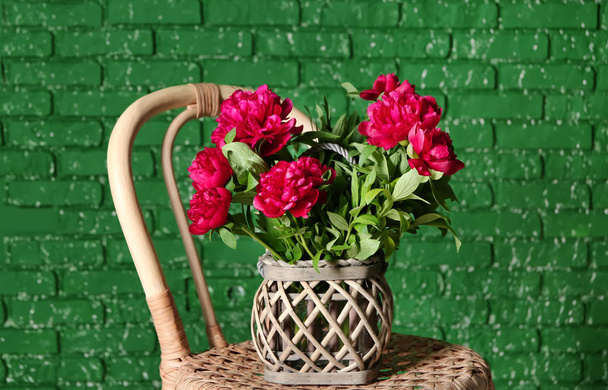 Vaas met mooie pioenroos bloemen op rieten stoel in de buurt van kleur bakstenen muur - Foto, afbeelding