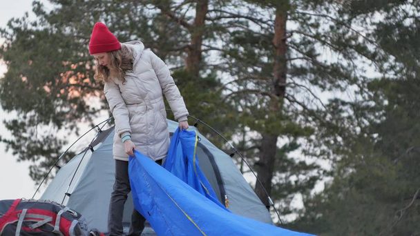 Привлекательная молодая туристка в красной шляпе собирает туристическую палатку возле леса на побережье
 - Фото, изображение