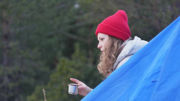 Приваблива молода жінка турист у червоній шапці сидить у туристичному наметі і п'є чай з термоса
 - Фото, зображення