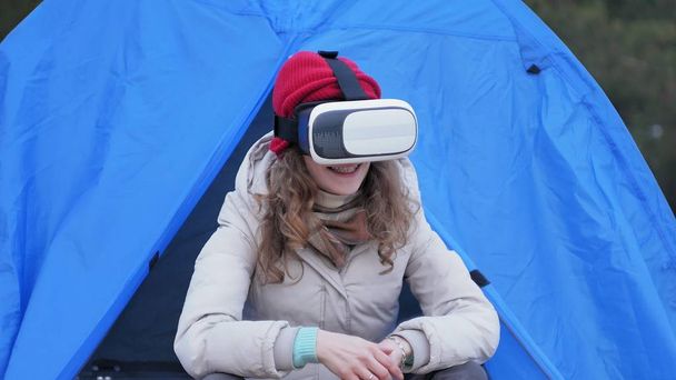 Attractive young woman tourist in a red hat sits in a tourist tent and enjoys a virtual reality helmet - Photo, Image