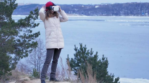 Приваблива молода жінка турист в червоному капелюсі сидить на березі і насолоджується шоломом віртуальної реальності
 - Фото, зображення
