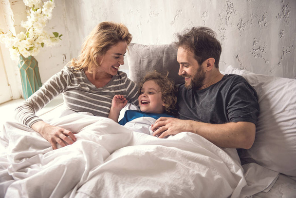 Cheerful parents are embracing son while staying in bed linen - Фото, изображение