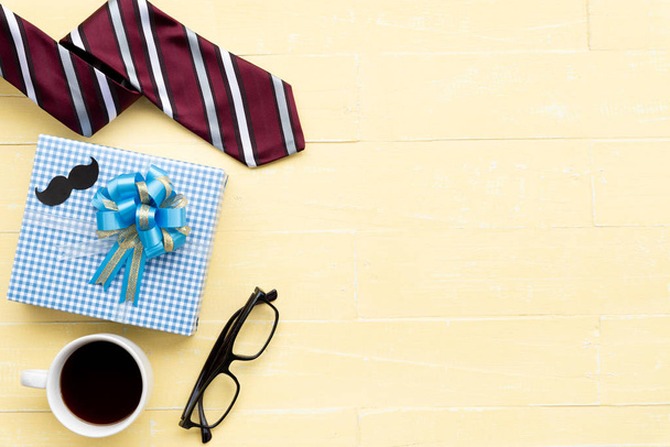 Happy fathers day concept. Red tie, glasses, gift box with mustache and coffee cup on bright yellow pastel wooden table background. - Photo, Image