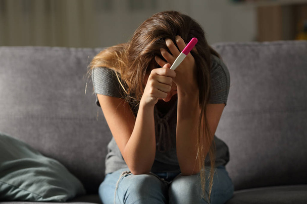 Single sad woman complaining holding a pregnancy test sitting on a couch in the living room at home - Photo, image