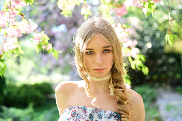 Calm blond girl enjoying spring day in floral garden filled with fresh aroma of flower blossom. Pretty young lady with blue eyes hiding in shade of blooming trees. Tranquility and mindfulness concept. - Foto, afbeelding