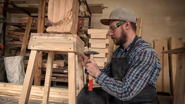 Πεπειραμένος ξυλουργός σε ρούχα εργασίας και μικρά buiness ιδιοκτήτης εργάζεται σε εργαστήριο ξυλουργικής, χρησιμοποιώντας ένα σφυρί, χτυπώντας τα καρφιά, στο τραπέζι είναι ένα σφυρί και πολλά εργαλεία - Φωτογραφία, εικόνα