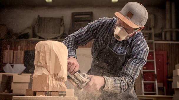 Ένας νεαρός άνδρας ξυλουργός σε ένα κοστούμι, γυαλιά πριονίζει το κεφάλι ενός άνδρα με ένα δέντρο, χρησιμοποιώντας μια γωνία αλεστικό μηχάνημα στο εργαστήριο, γύρω από πολλά εργαλεία για την εργασία - Φωτογραφία, εικόνα