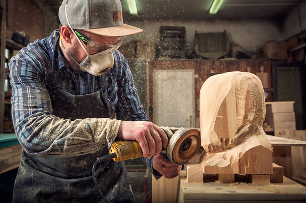 carpinteiro experiente em roupas de trabalho serra a cabeça de um homem com uma árvore, usando um moedor de ângulo na oficina, em torno de um monte de ferramentas para o trabalho
 - Foto, Imagem