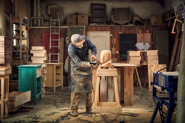 Carpinteiro, pequeno dono de buiness em roupas de trabalho serra a cabeça de um homem com uma árvore, usando um moedor de ângulo na oficina, em torno de um monte de ferramentas, de madeira, móveis para o trabalho
 - Foto, Imagem