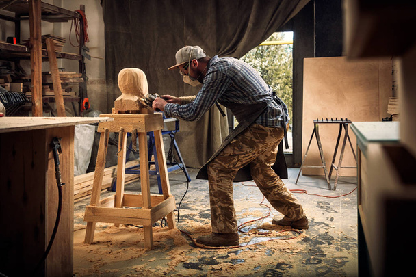 Carpintero, pequeño propietario de la ropa de trabajo sierra para cortar la escultura de madera de la cabeza de un hombre, utilizando una amoladora angular en el taller, alrededor de una gran cantidad de herramientas, madera, muebles para el trabajo
  - Foto, imagen