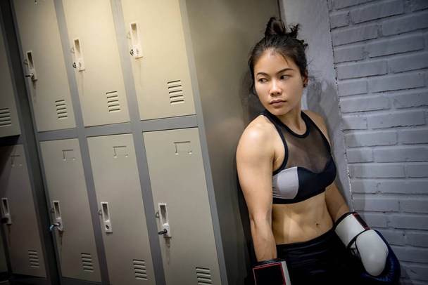 Joven boxeadora asiática con guantes de boxeo de pie junto al armario en el vestuario
. - Foto, Imagen